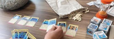 Photographie montrant une partie de jeu de cartes &eacute;ducatif. Plusieurs cartes dispos&eacute;es en rang&eacute;e et un sablier orange en haut &agrave; droite. Main humaine en train de manipuler une carte, plusieurs paquets de cartes empil&eacute;s &agrave; droite, et un sac en tissu beige contenant des jetons en forme de lettres.