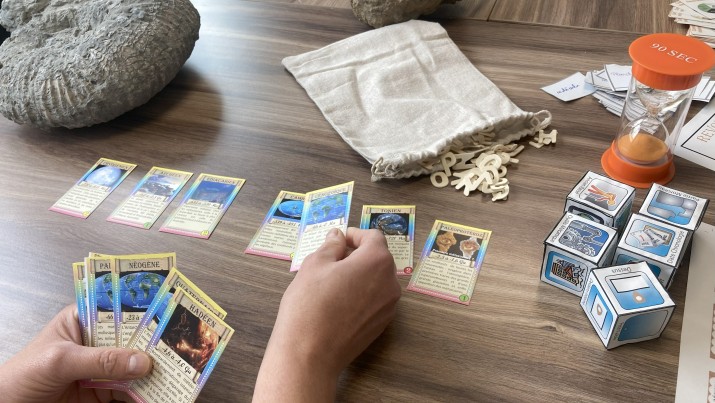 Photographie montrant une partie de jeu de cartes &eacute;ducatif. Plusieurs cartes dispos&eacute;es en rang&eacute;e et un sablier orange en haut &agrave; droite. Main humaine en train de manipuler une carte, plusieurs paquets de cartes empil&eacute;s &agrave; droite, et un sac en tissu beige contenant des jetons en forme de lettres.
